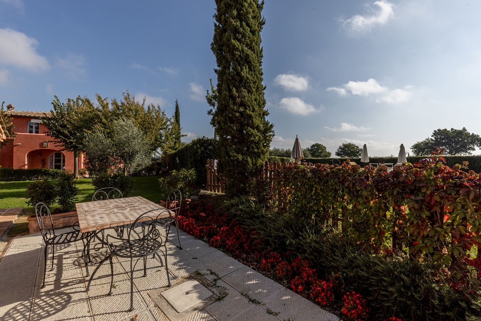 Un pati amb taula i cadires, envoltat d'un jardí ple de flors vermelles i un arbre alt.