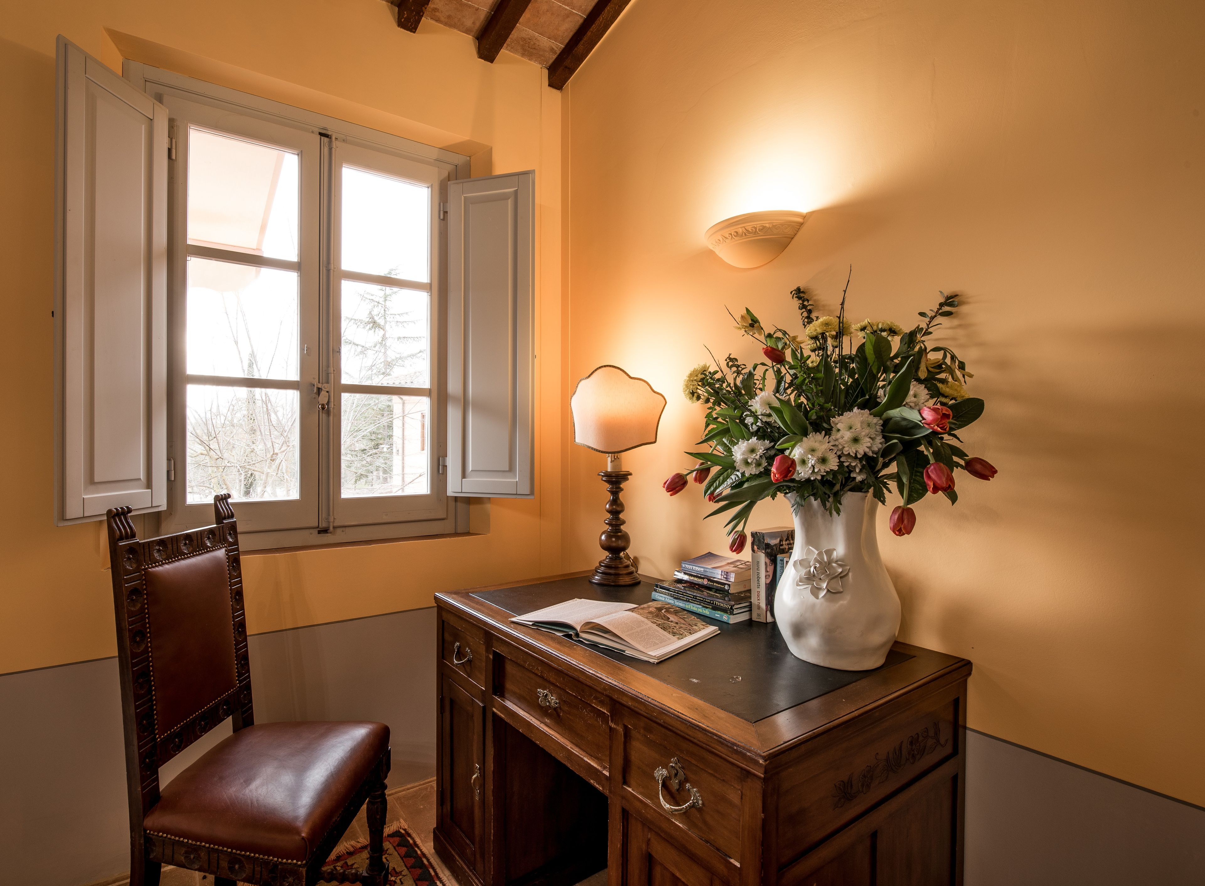 A wooden desk with a vase of flowers, a lamp, and books.