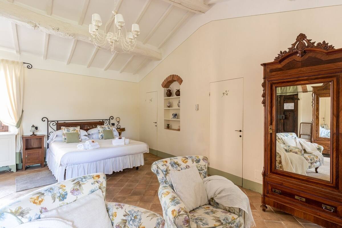A bedroom with a large bed, a wooden wardrobe, a floral armchair, a wooden dresser, and a mirror.