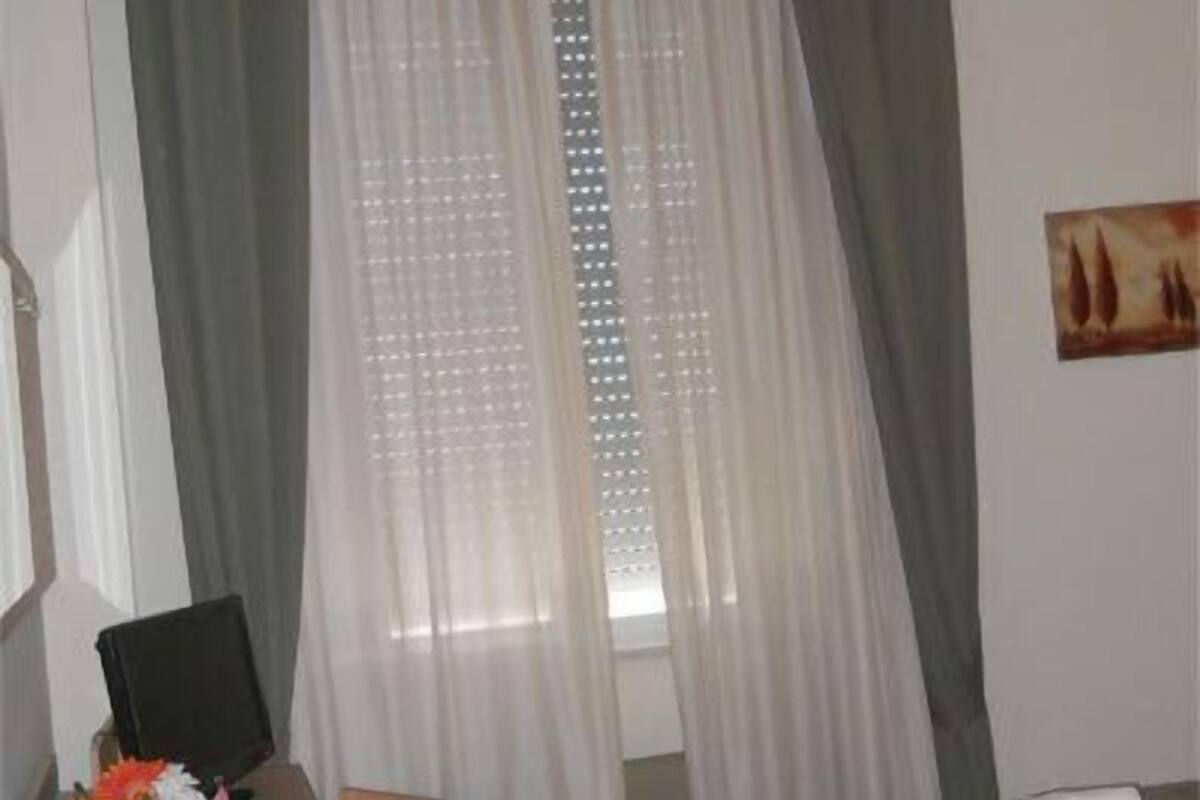Una habitación de hotel con cama, escritorio con silla y ventana con cortinas traslúcidas.