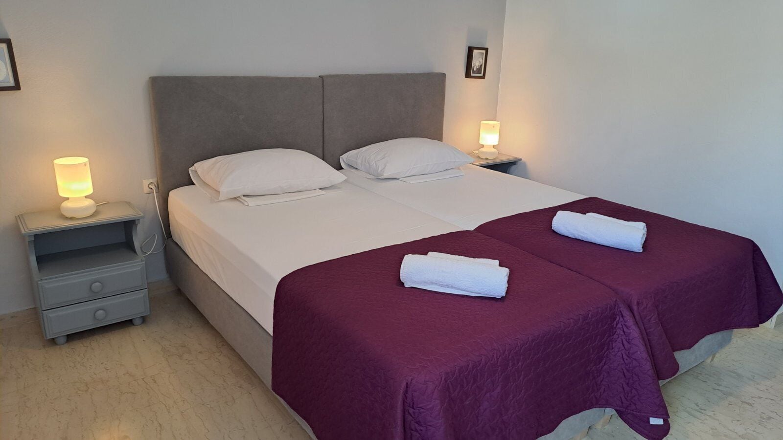 A double bed with two white pillows, a grey headboard, and a purple bedspread. There are two bedside tables with lamps, one on each side of the bed.