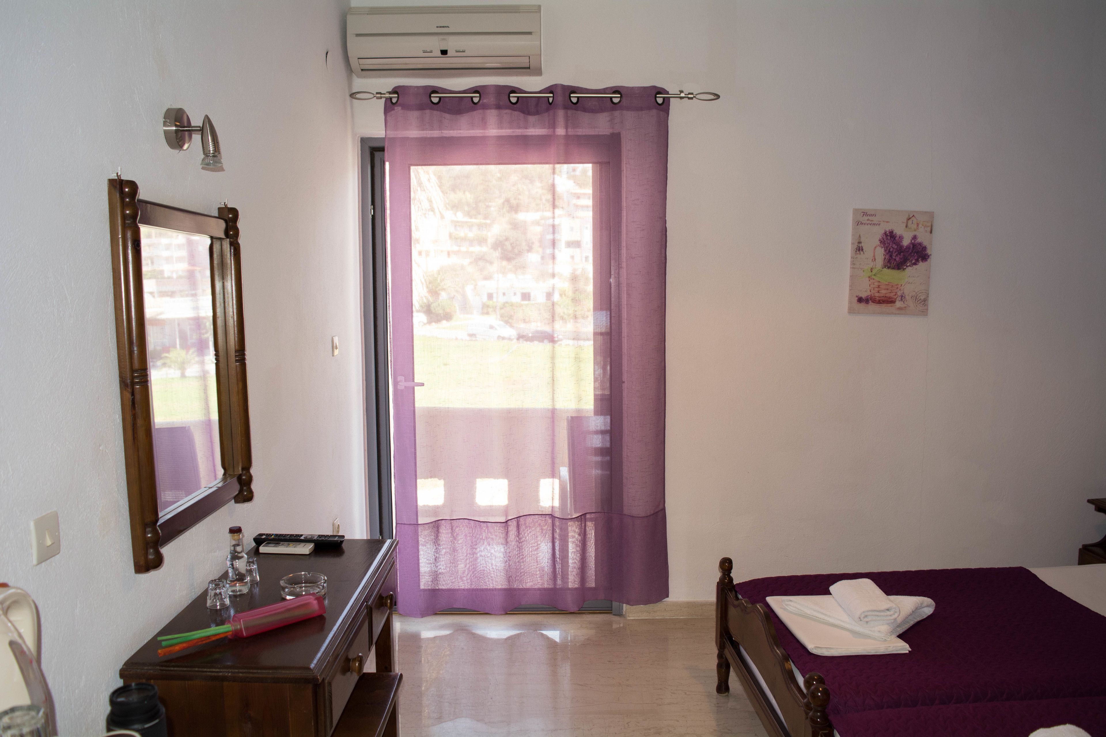A hotel room with a bed, a desk with a mirror, a window with purple curtains, and a small picture on the wall.