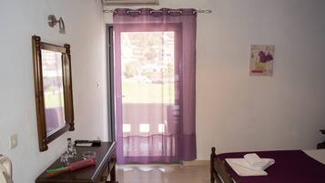 A hotel room with a bed, a desk with a mirror, a window with purple curtains, and a small picture on the wall.