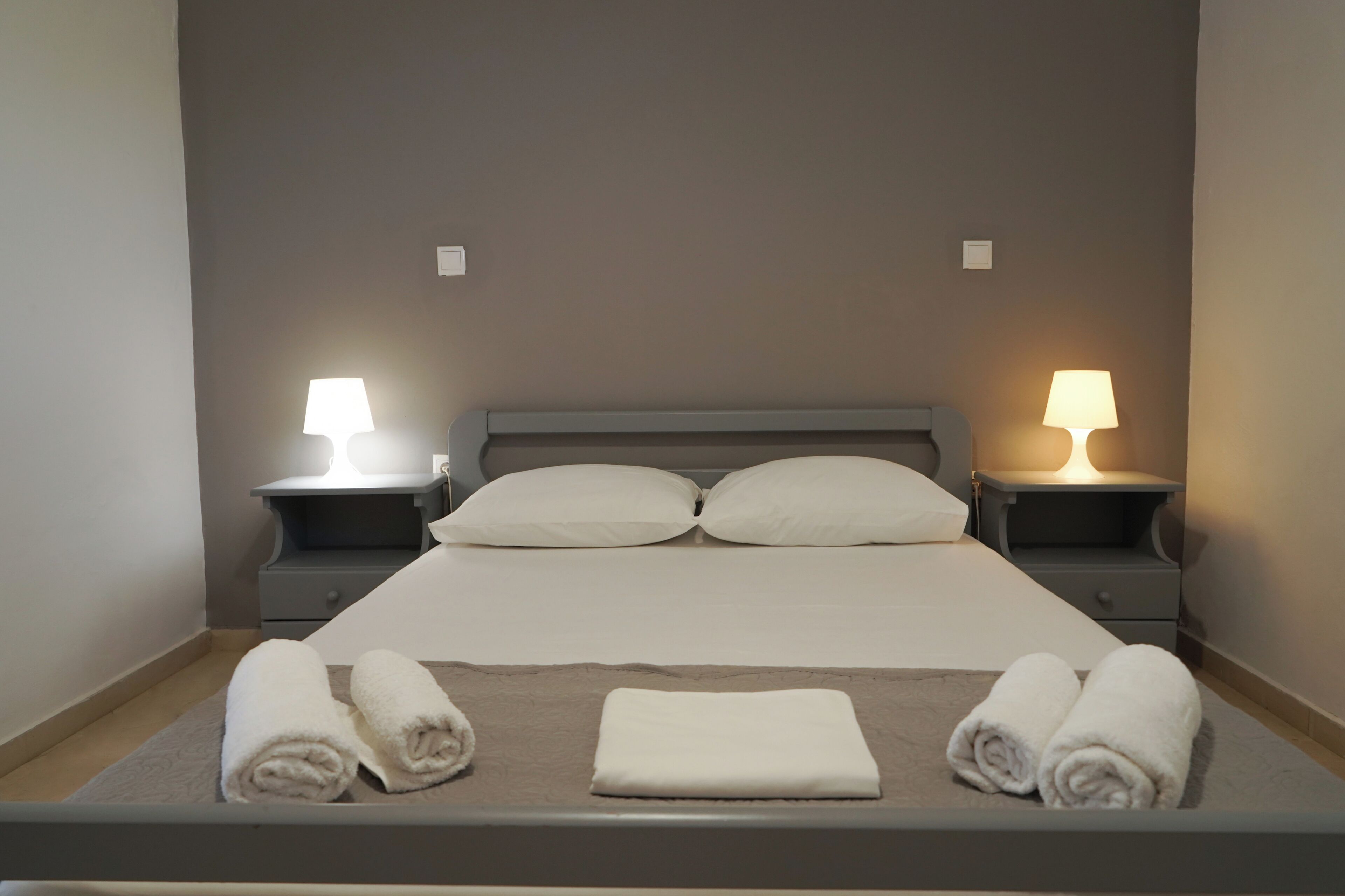 A neatly made bed with two pillows, two bedside lamps, and three rolled towels on the bed.