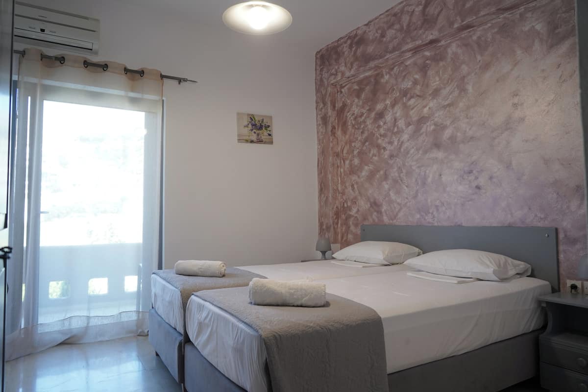 Un dormitorio con dos camas, ventilador de techo, mesita de noche y ventana con cortinas traslúcidas.