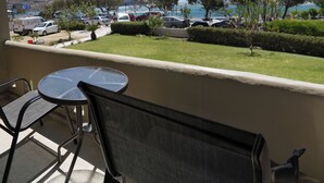 A balcony with a table and chairs, overlooking a parking area with cars and a view of the sea and mountains. - Galini Beach (Agios Vasileios)