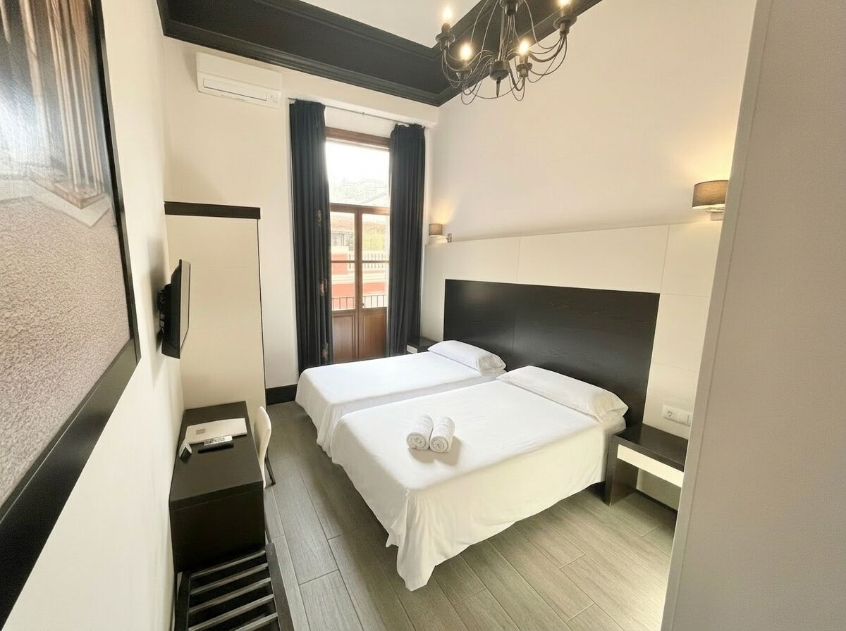 A hotel room with two beds, a desk, a chair, a window with curtains, and a chandelier.