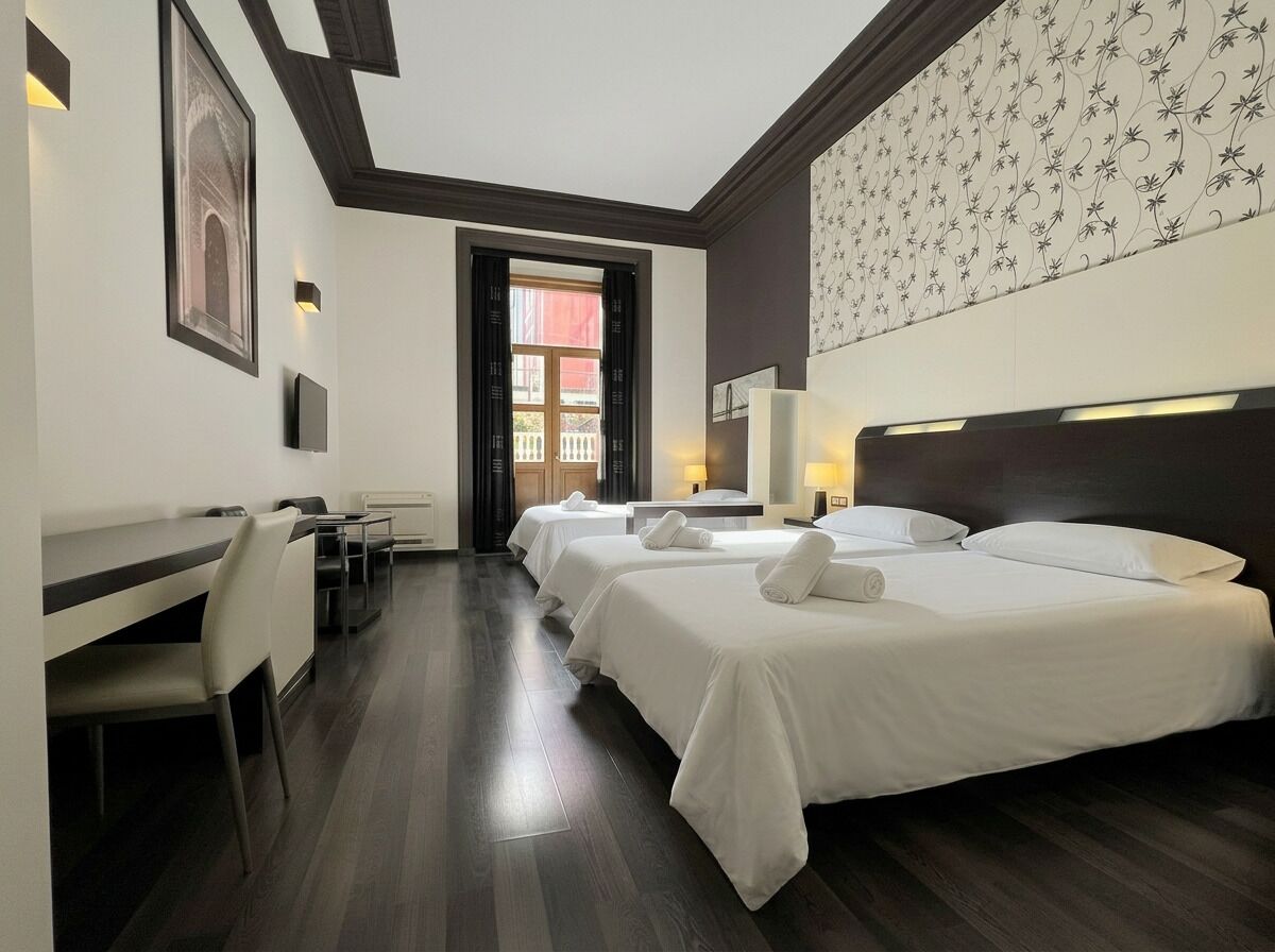 Une chambre d’hôtel avec un grand lit, un bureau et une chaise. Il y a une fenêtre avec des rideaux, un mur orné d’un motif floral et un tableau encadré accroché au mur.
