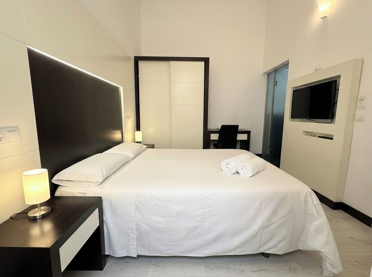 A hotel room with a bed, bedside table, lamp, TV, and a wardrobe.