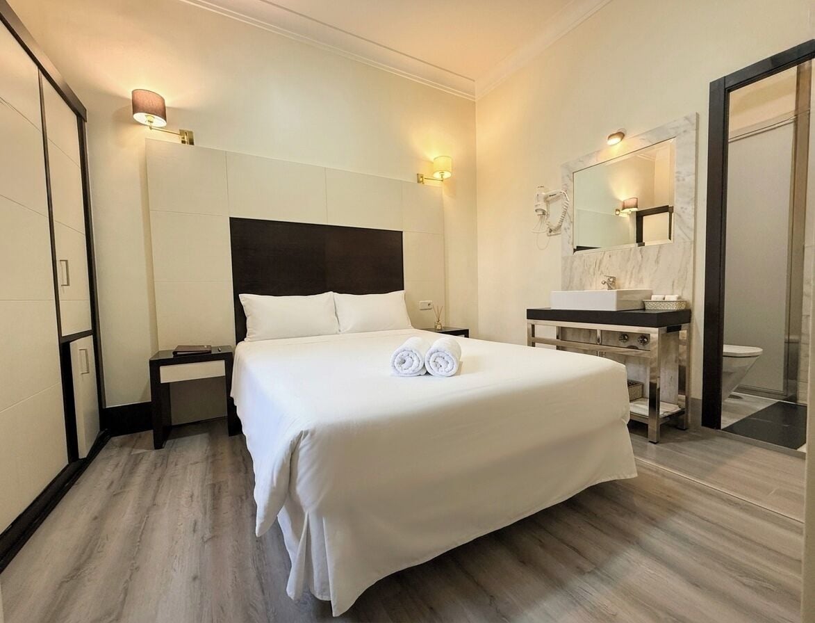 Una habitación de hotel con una cama grande, mesitas de noche, un baño con espejo y lavamanos.