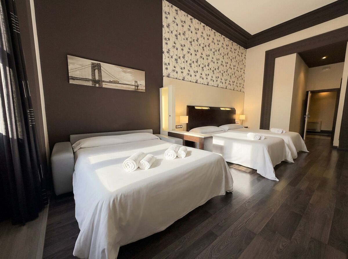 A hotel room with three beds, a headboard with integrated lighting, a large window, and a wall-mounted artwork.