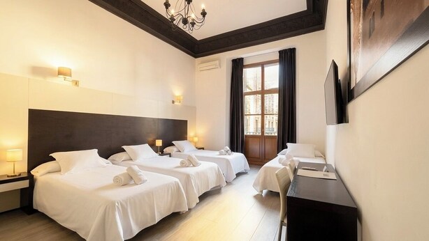Une chambre d’hôtel avec trois lits, un bureau, un lustre et une grande fenêtre avec des rideaux.