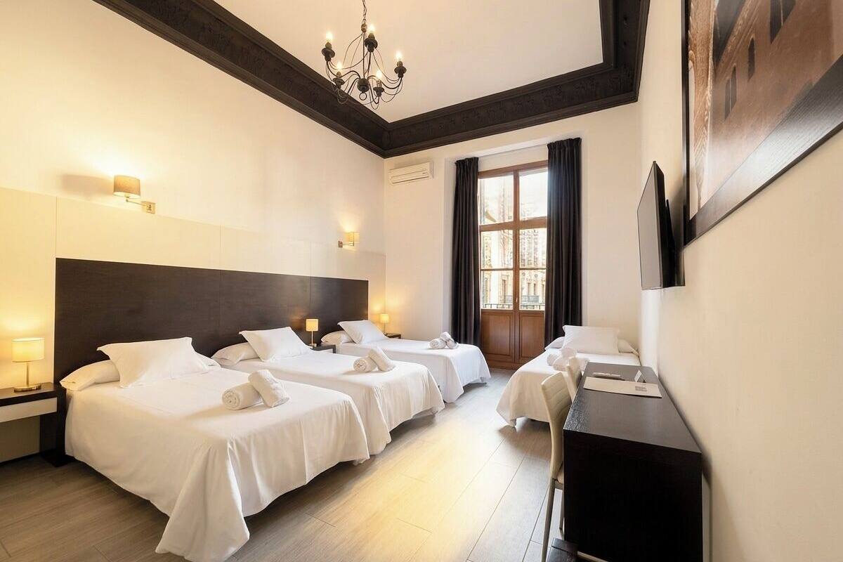 Habitación de hotel con tres camas, un escritorio, una araña y un amplio ventanal con cortinas.