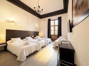 Quarto de hotel com três camas, uma escrivaninha, um candelabro e uma janela grande com cortinas.