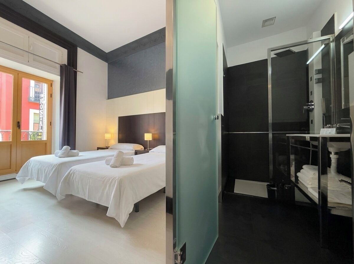 A hotel room with two beds, a bathroom with a glass shower, and a view of a building outside.