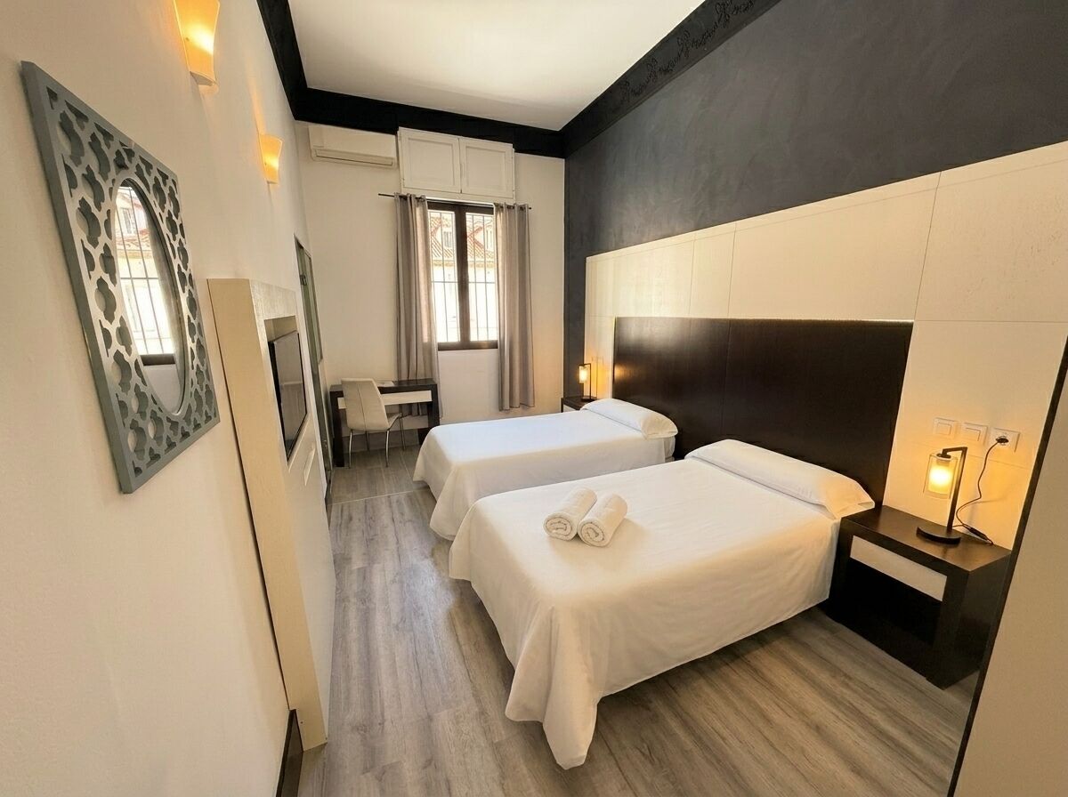 A hotel room with two beds, a large mirror, a bedside table with a lamp, and a window with curtains.
