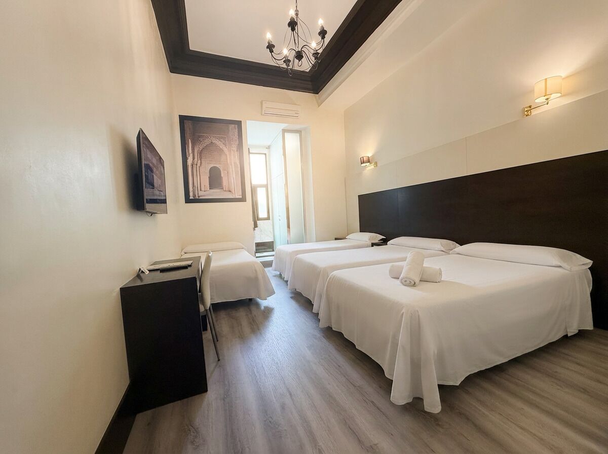 A hotel room with three beds, a desk, a chair, a chandelier, and a painting on the wall.