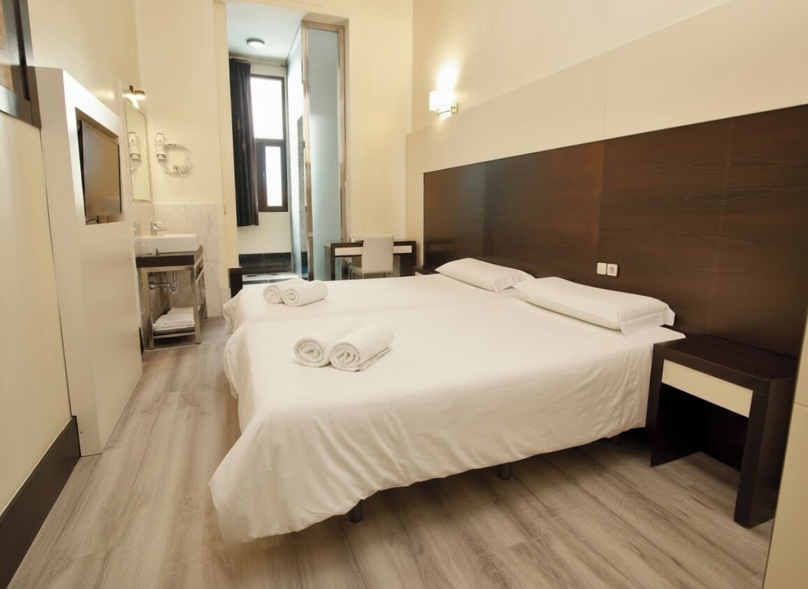 A hotel room with a large bed, a bedside table, a mirror, a small desk, and a bathroom with a sink and mirror.