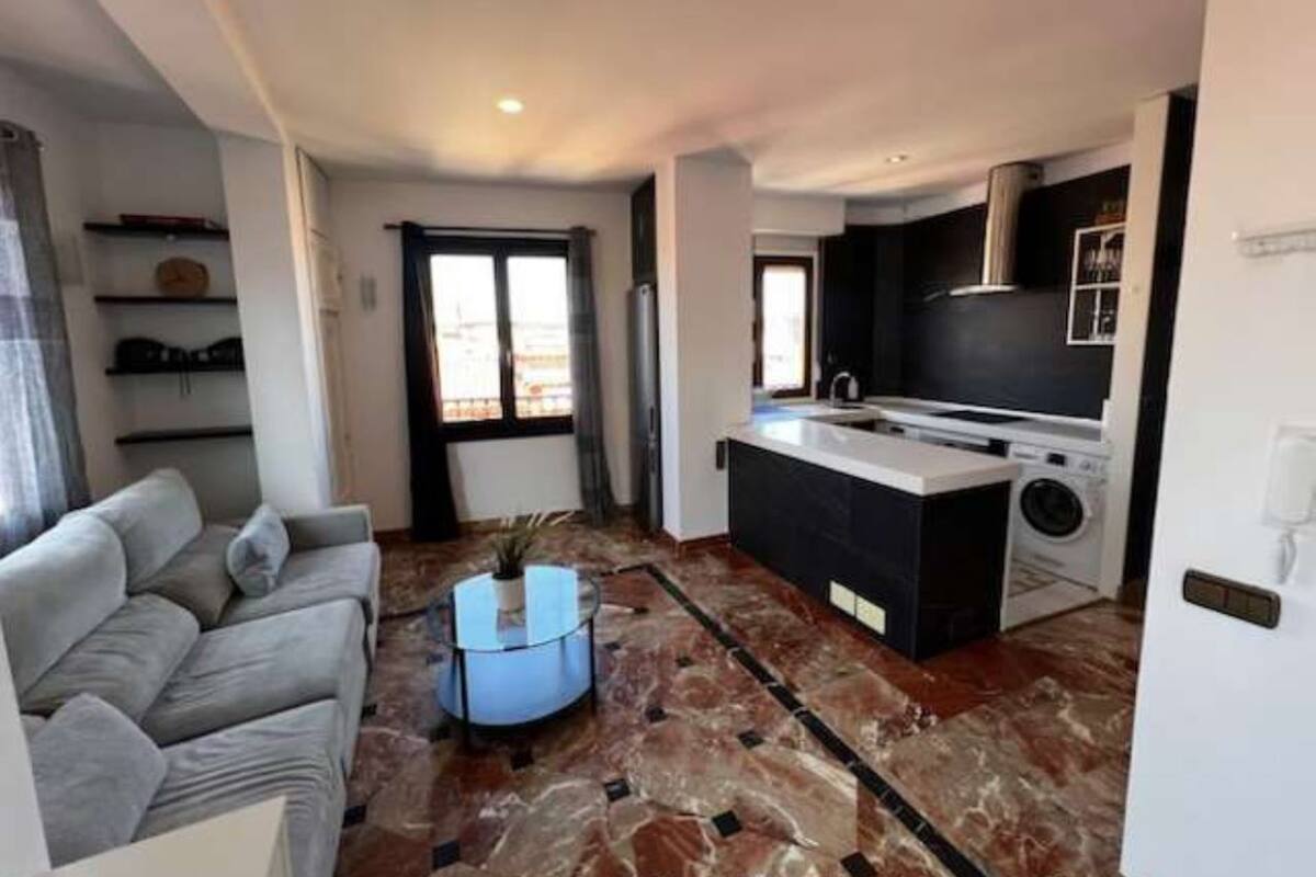 Una sala moderna con un sofá gris, una mesita de centro y una zona de cocina con televisor, lavadora y una pequeña isla.