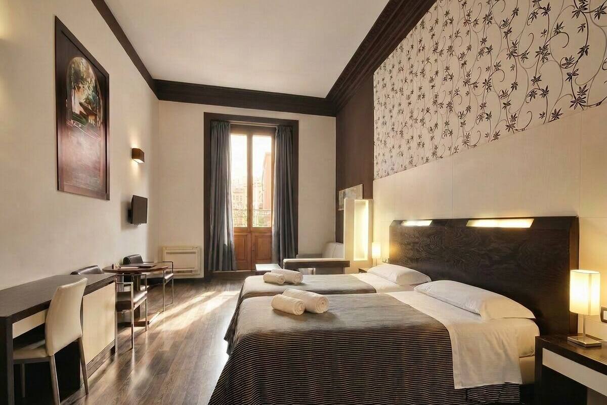 Habitación de hotel con una cama grande, un escritorio y una silla. Se ve una ventana con cortinas, un televisor de pared y un adorno mural con motivos.
