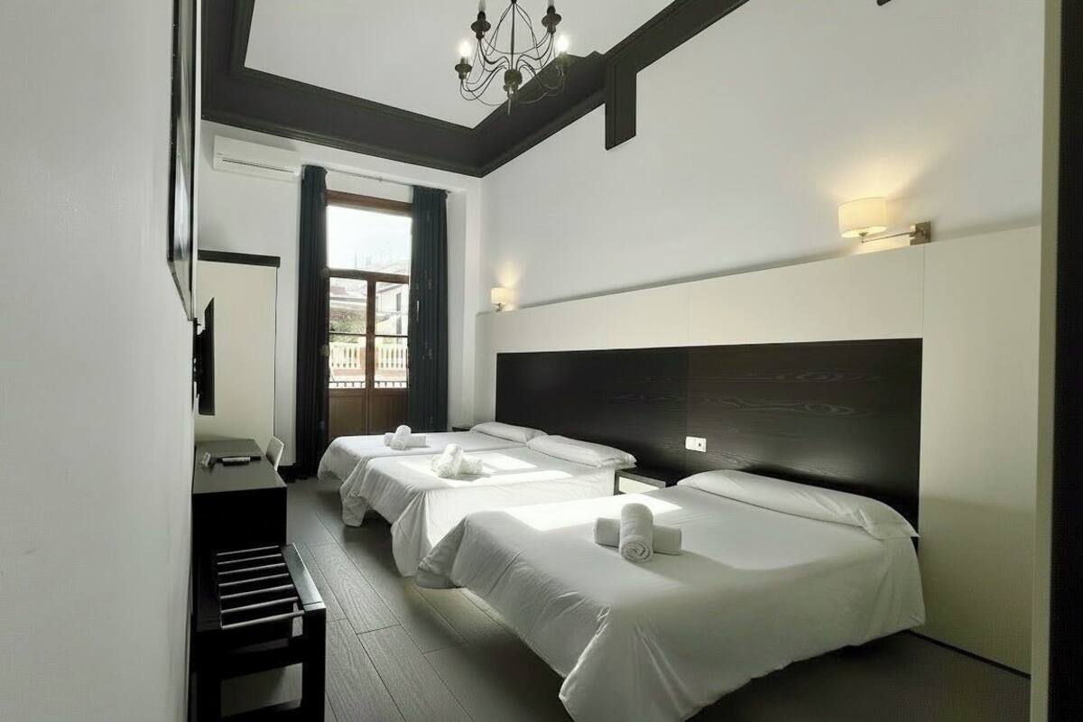 Habitación de hotel con dos camas, un escritorio, una silla, una araña y una ventana con cortinas.