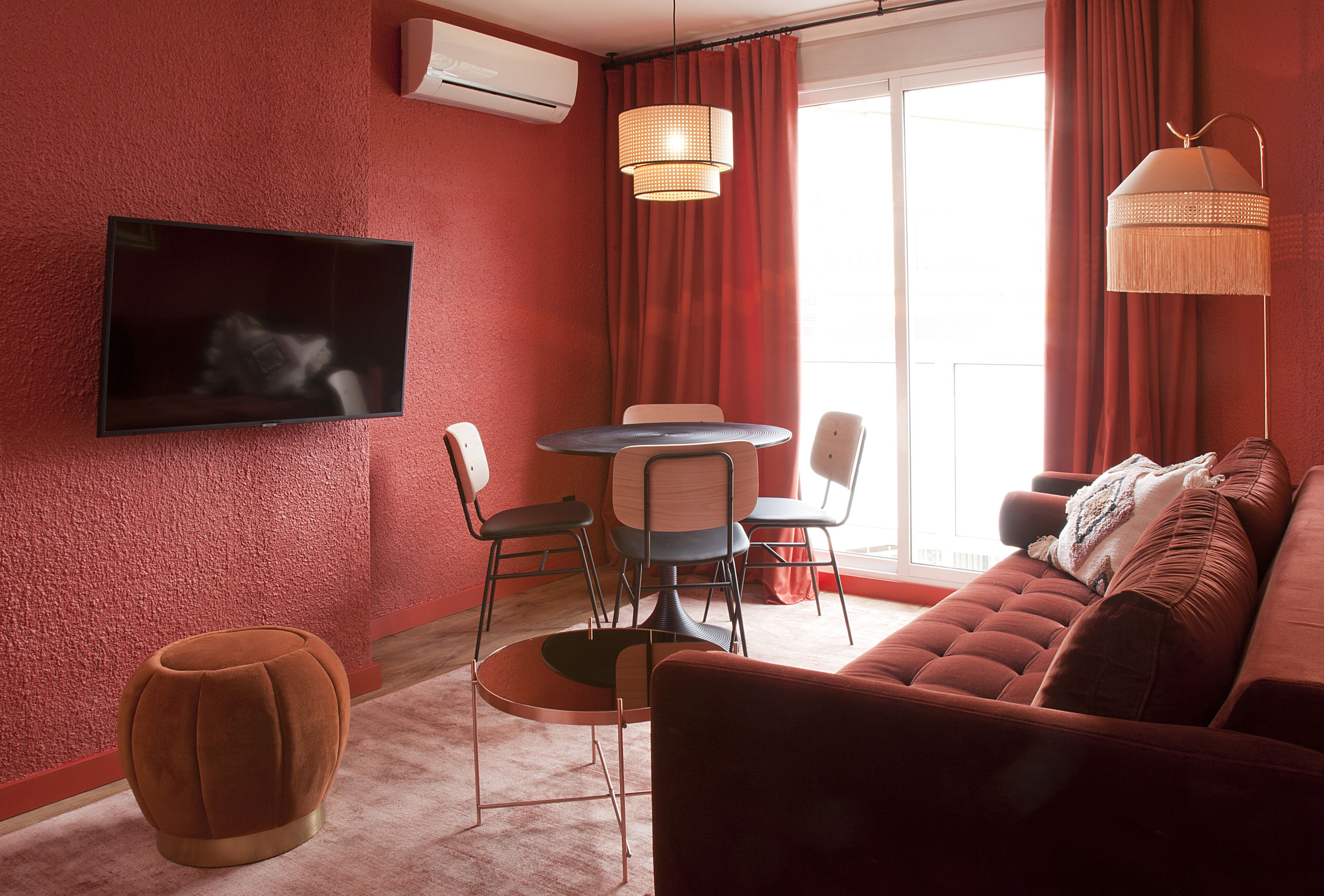 A modern living room with a red wall, a flat-screen TV mounted above a sofa, a dining area with chairs and a table, and a large window with curtains.