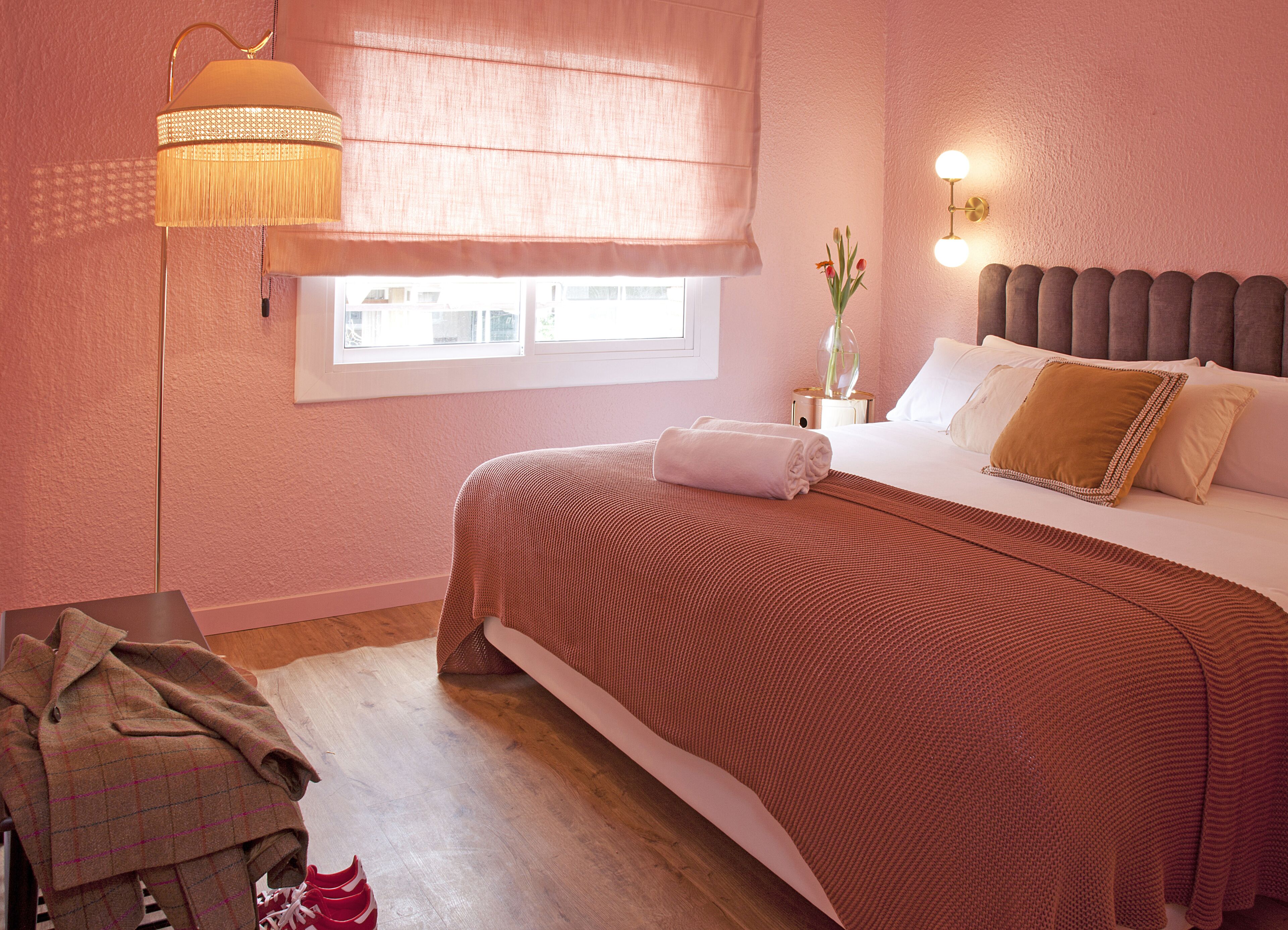 A bedroom with a large bed, a bedside table with a lamp, a window with blinds, and a view of buildings outside.