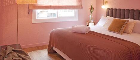 A bedroom with a large bed, a bedside table with a lamp, a window with blinds, and a view of buildings outside.