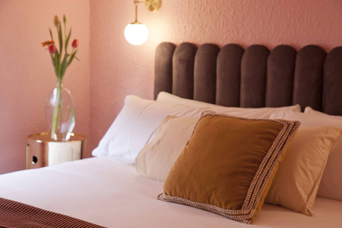A neatly made bed with white and brown pillows, a clear vase with flowers, and a wall-mounted light fixture.