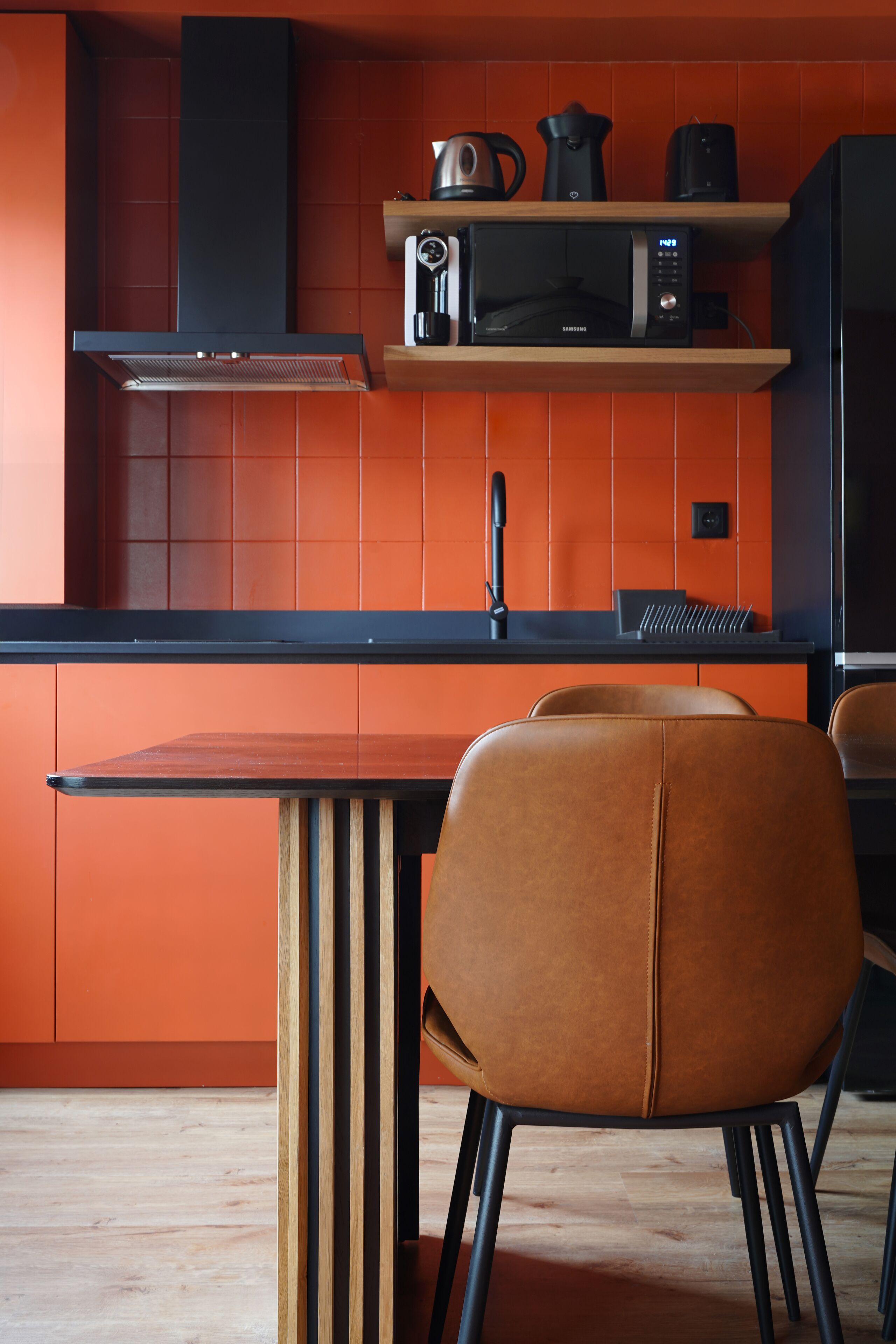 A modern kitchen with orange tiles, a black microwave, and a wooden table with two chairs.