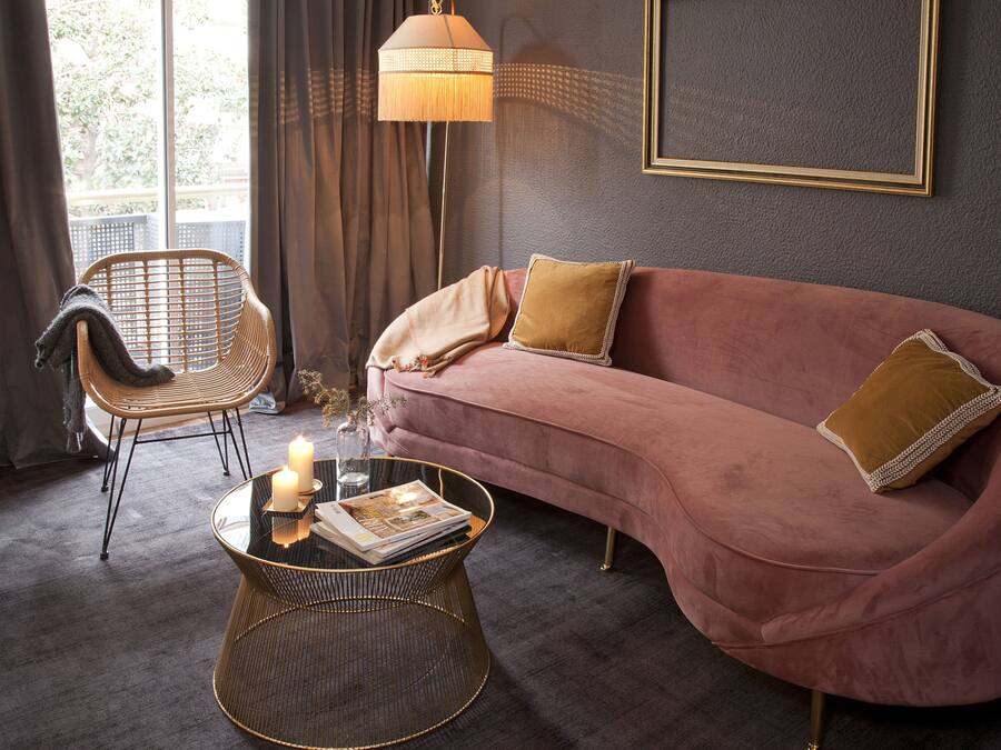 Una habitación con un sofá rosa, una silla de mimbre, una mesa redonda de centro con velas y una lámpara de pie.