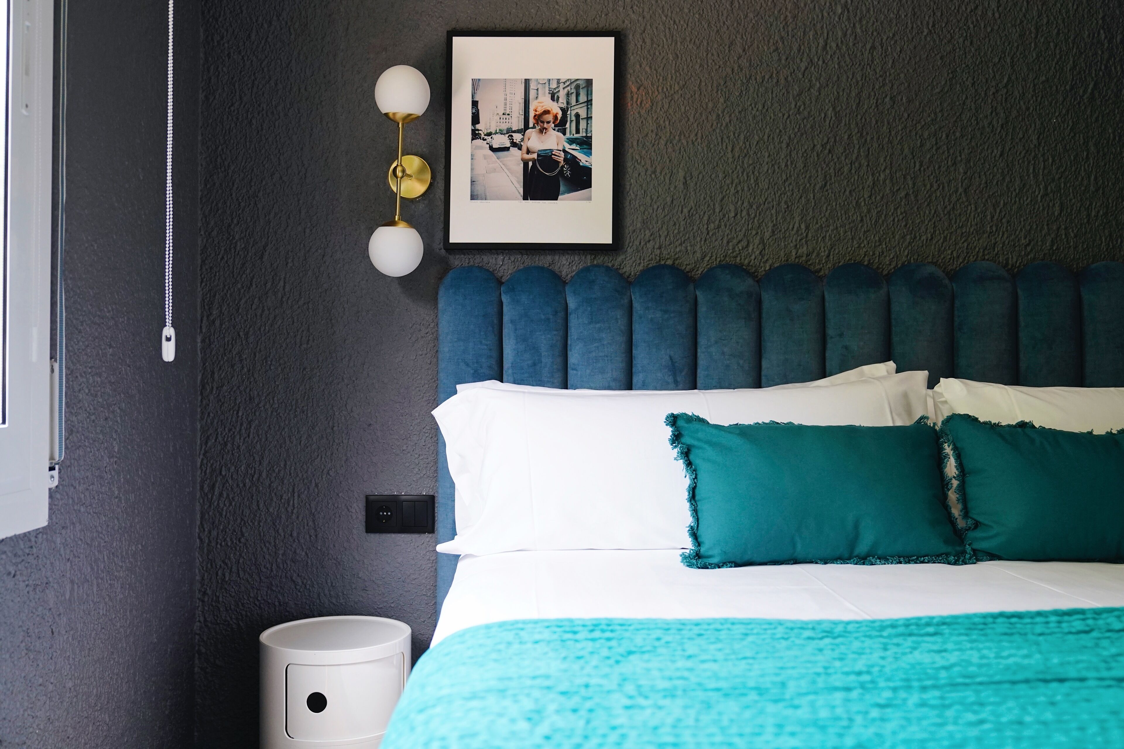 Un lit bien fait, avec une literie blanche et turquoise, un mur foncé à la texture intéressante et une photo encadrée au-dessus du lit.