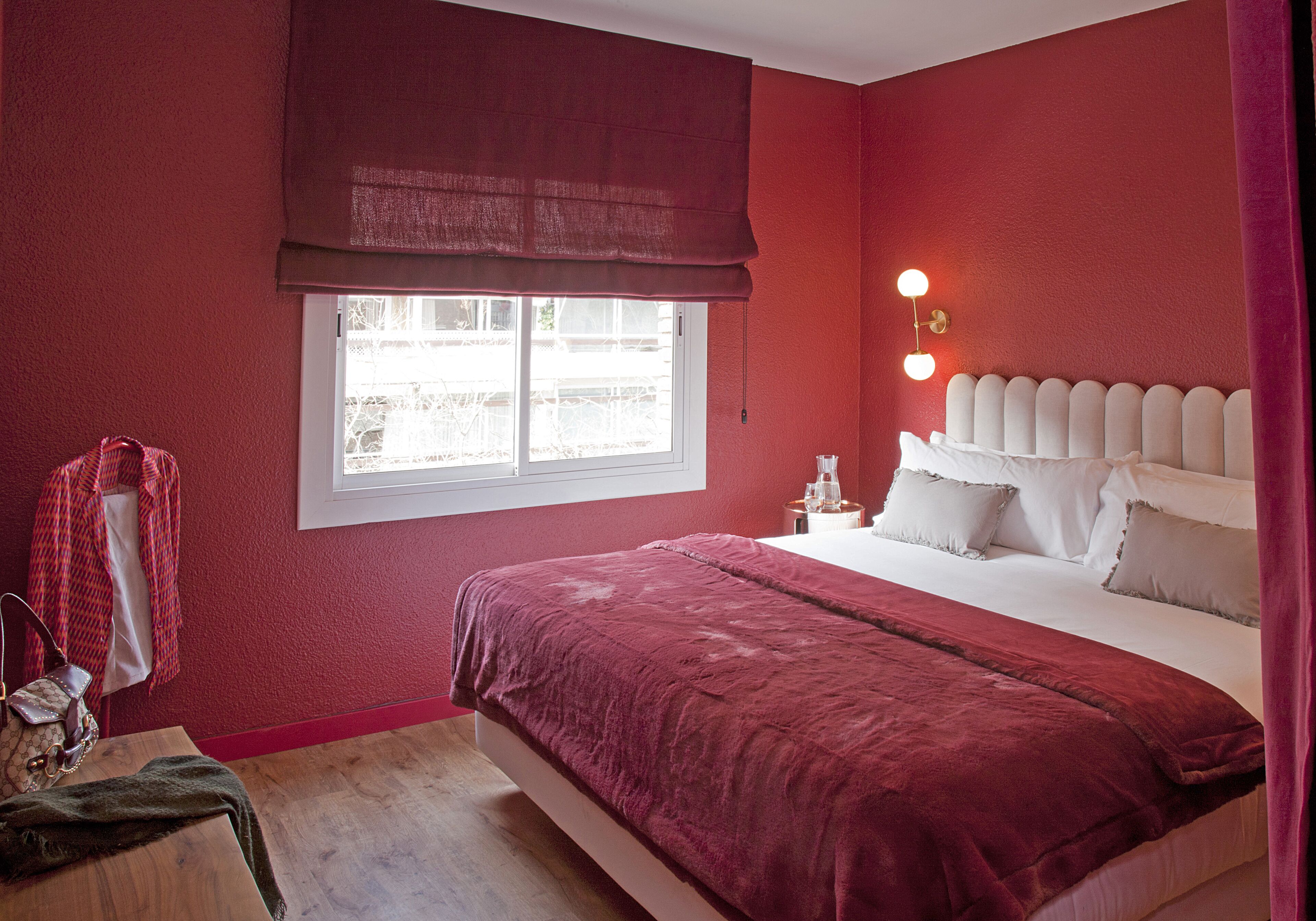 A bedroom with a large bed, a window with a red blind, a bedside table with a lamp, and a red wall.