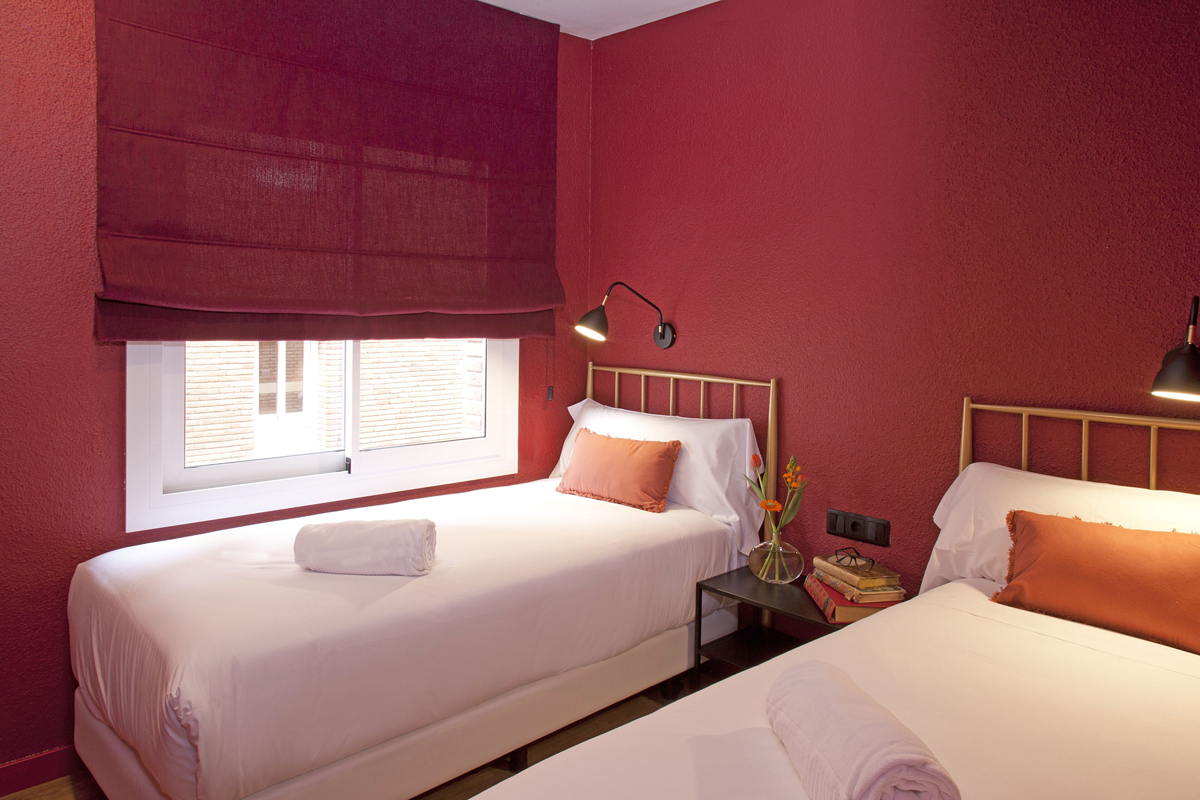 A hotel room with two beds, a window with blinds, a bedside table with a lamp, and a nightstand with books and a vase of flowers.