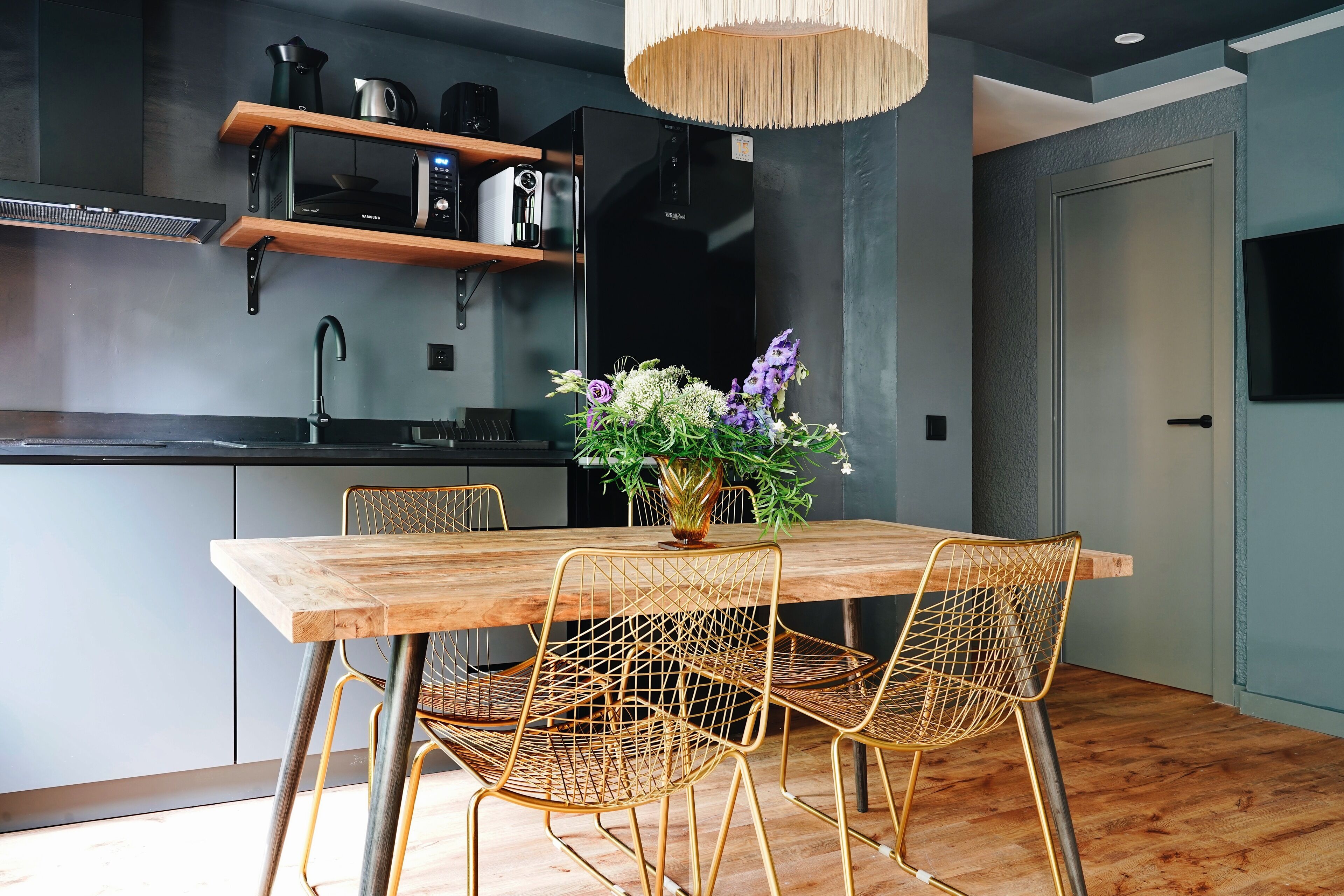 A modern kitchen with a wooden dining table, wire mesh chairs, and a vase of flowers.