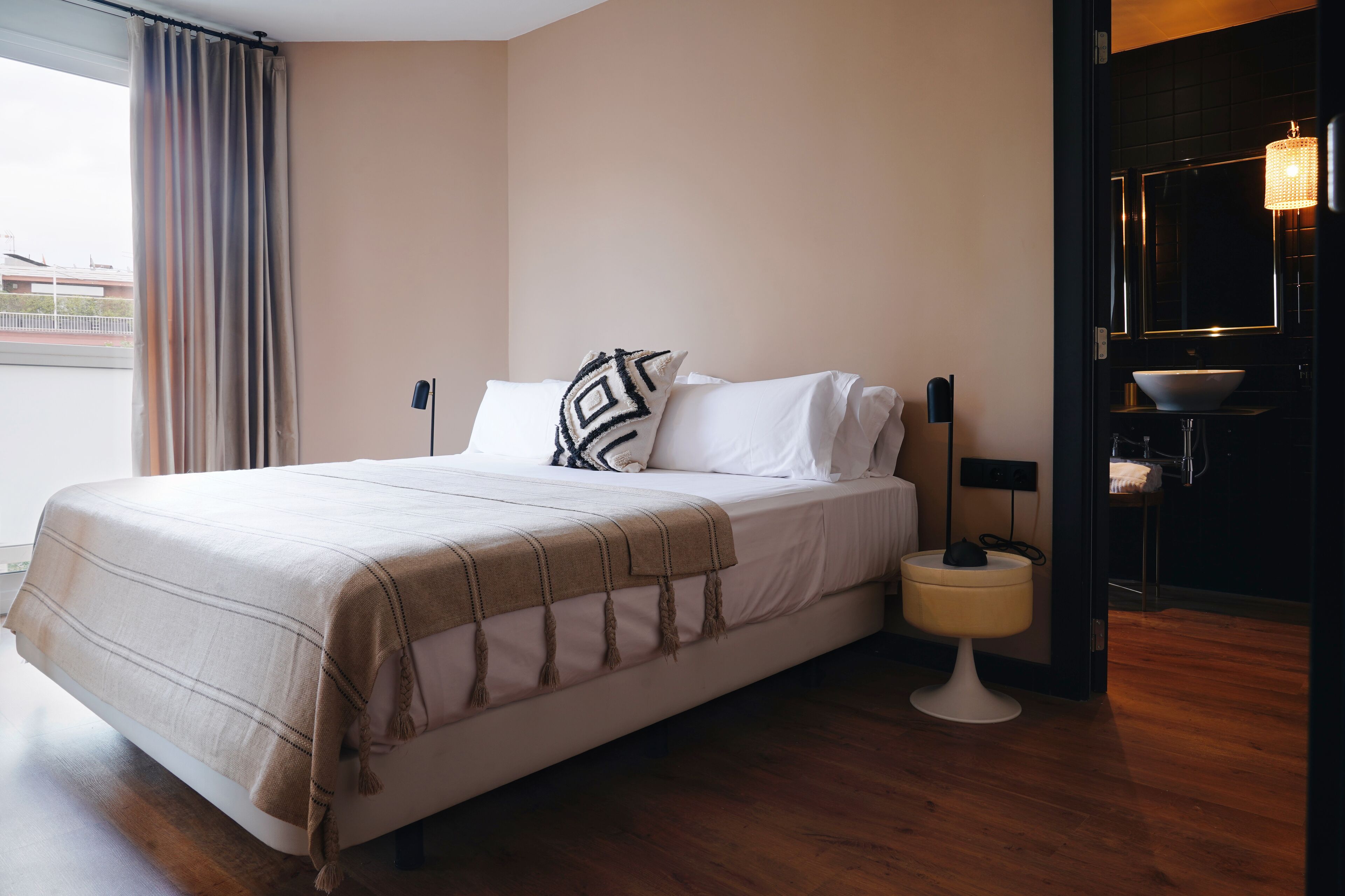 A neatly made bed with a beige blanket, a bedside table with a lamp, and a view of the outside through a window with curtains.