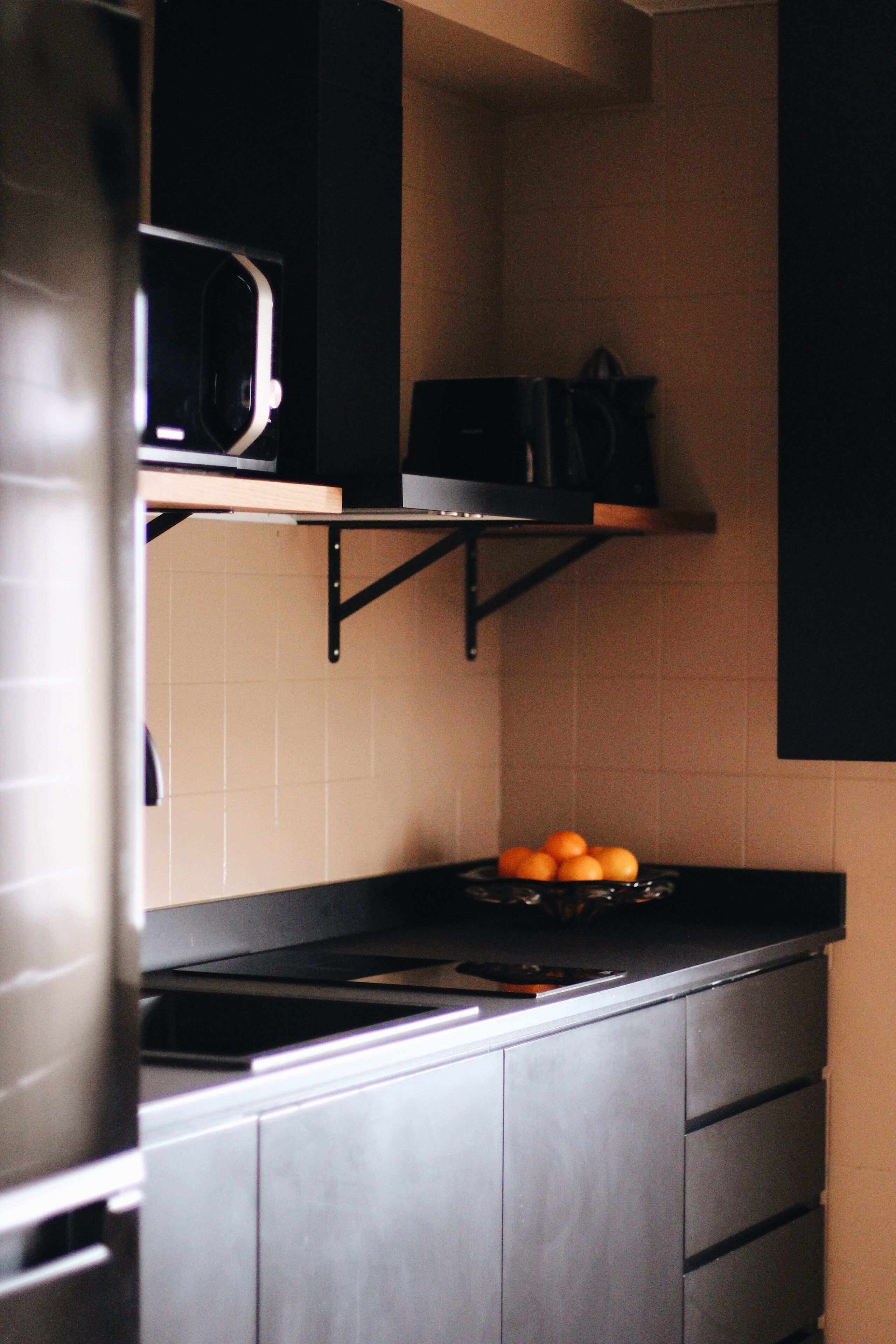 A kitchen with a black countertop, a microwave, and a shelf with oranges.