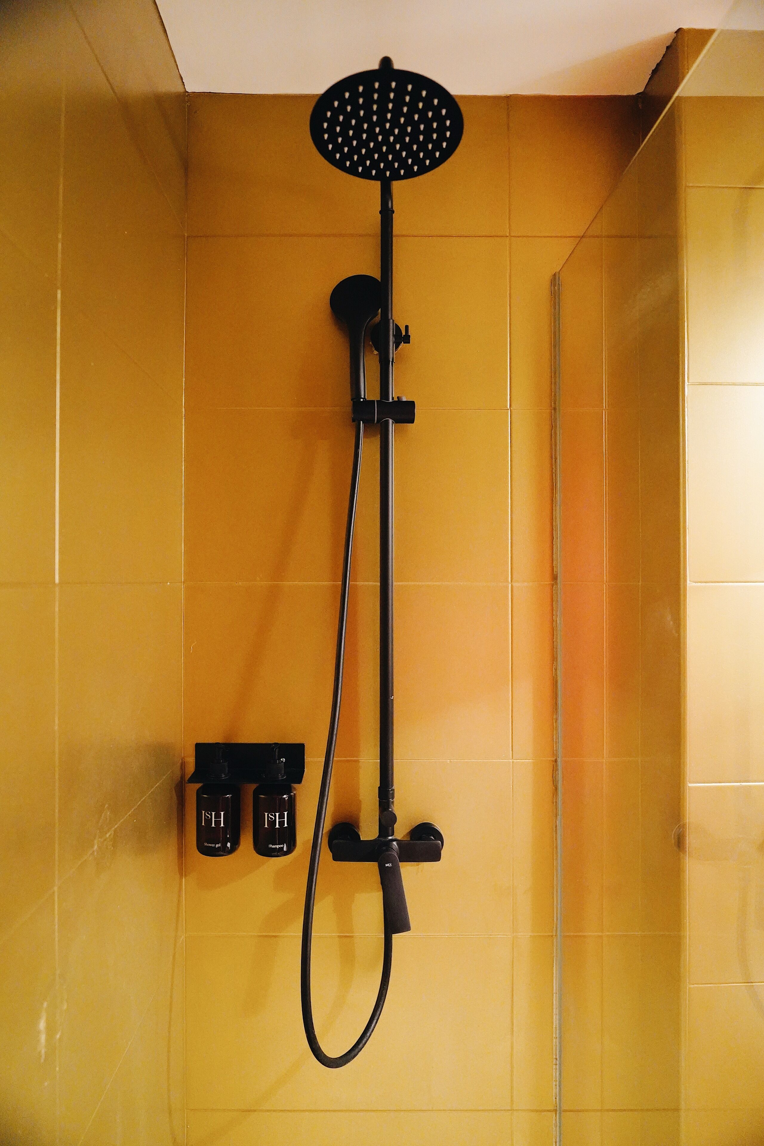 Ducha con cabezal de ducha manual y manguera larga, sobre un muro cubierto de azulejos amarillos.