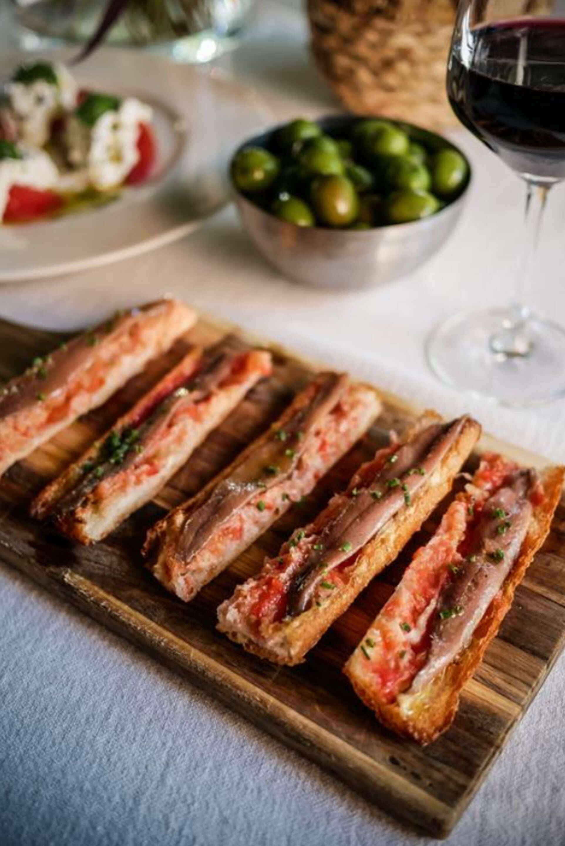 Una tabla de madera con rebanadas de carne a la parrilla, un cuenco de aceitunas y una copa de vino tinto sobre una mesa.