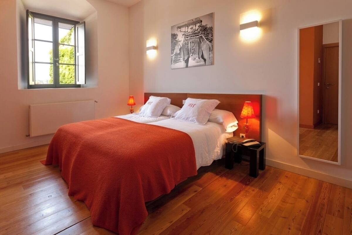 Ein Schlafzimmer mit Holzboden, einem Bett mit roter Decke, zwei Nachttischlampen, einem Nachttisch, einem Fenster mit Blick auf Grünflächen und einem Wandbild.