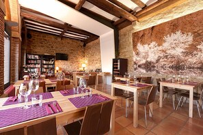 Un restaurant aux murs de pierre, aux poutres apparentes et aux tables décorées de sets de table violets et de verres à vin.