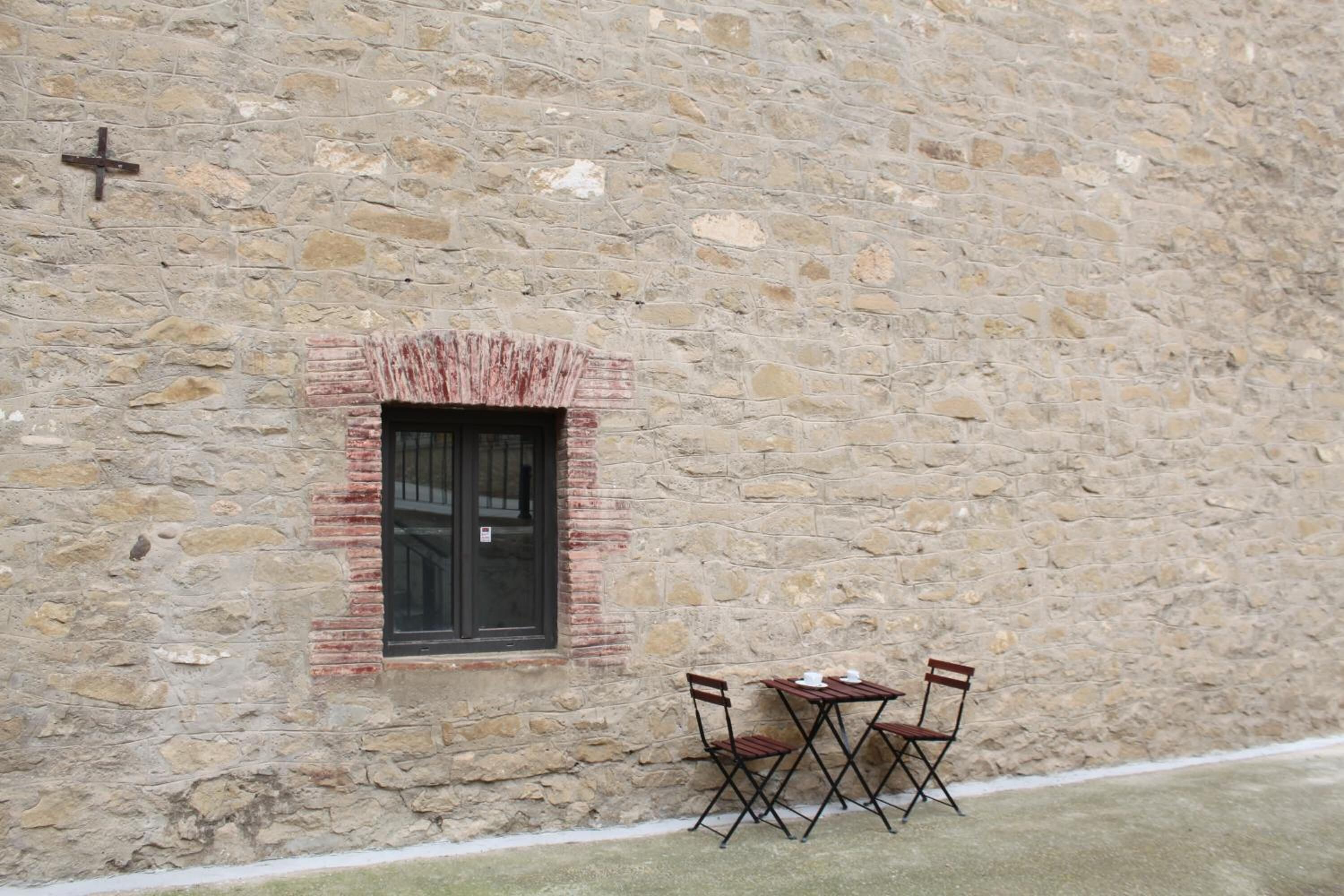 A stone building with a small window and a cross on the wall. There are two chairs and a table outside the window.