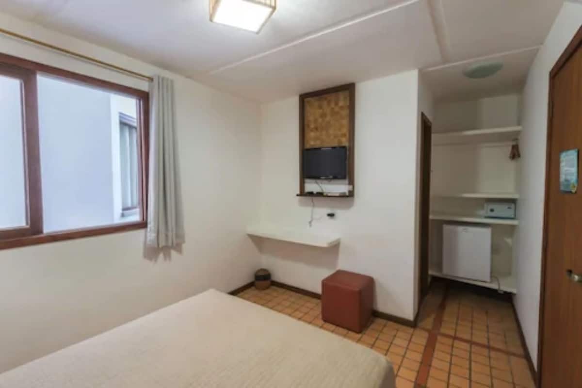 Quarto pequeno e limpo, com uma cama, televisão na parede, um banquinho pequeno e uma escrivaninha com computador.