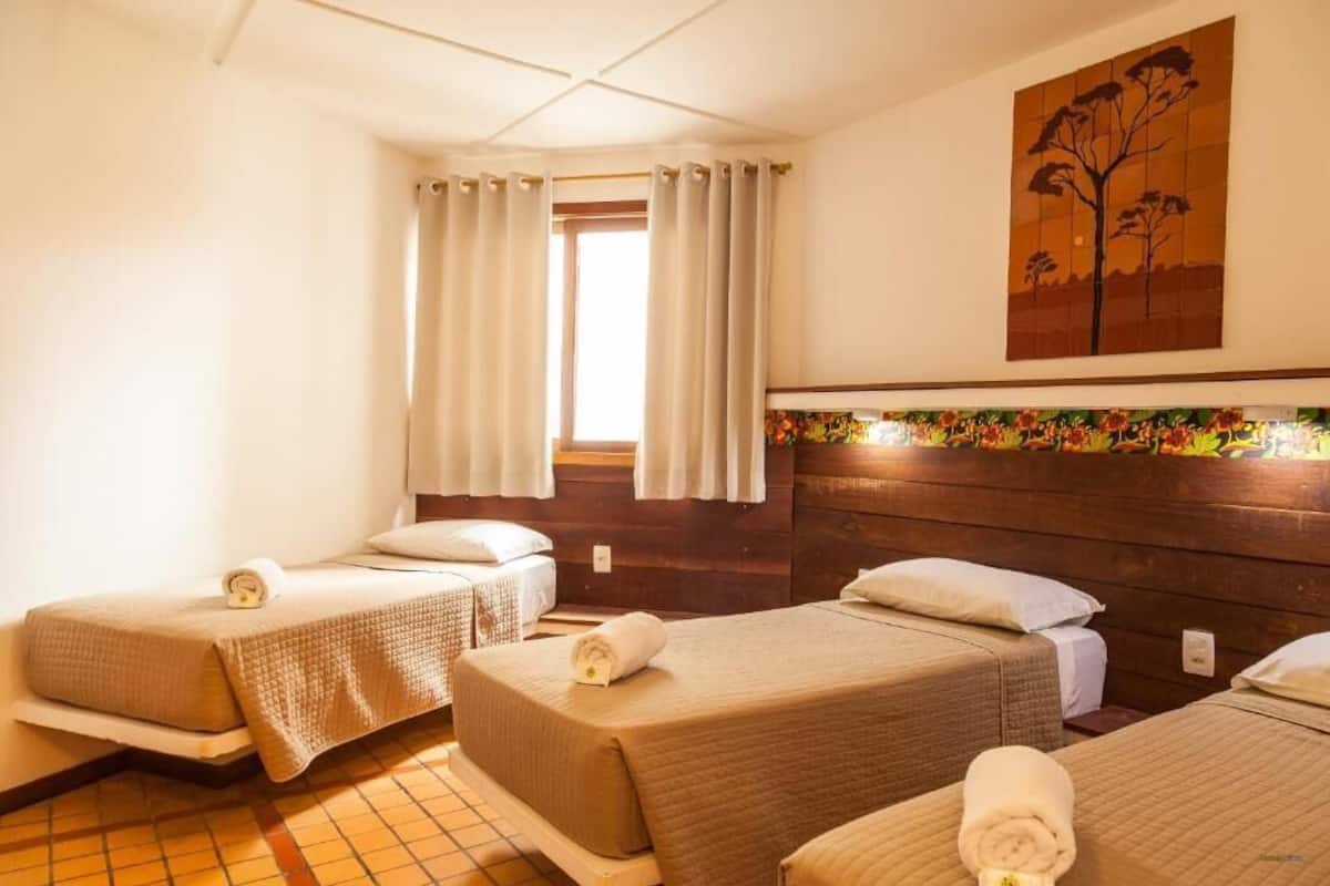 Quarto de hotel com duas camas, janela com cortinas, cabeceira de madeira e uma pintura de uma árvore.