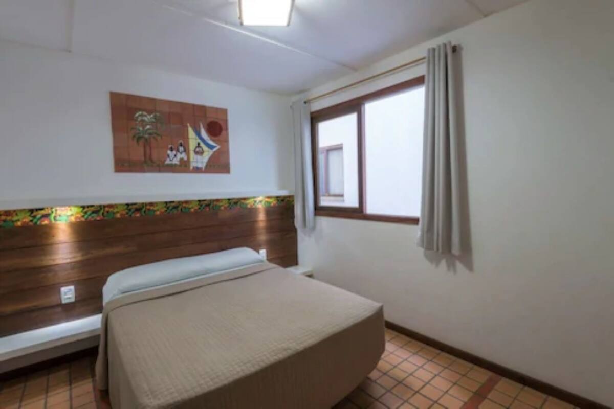 Quarto com cama, janela com cortinas, quadro na parede e chão ladrilhado.