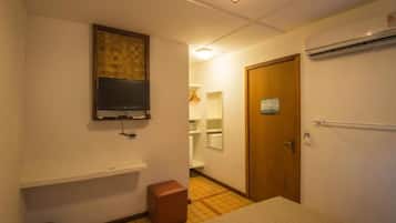 A small, compact hotel room with a single bed, a small stool, a television mounted on the wall, and a wooden door.