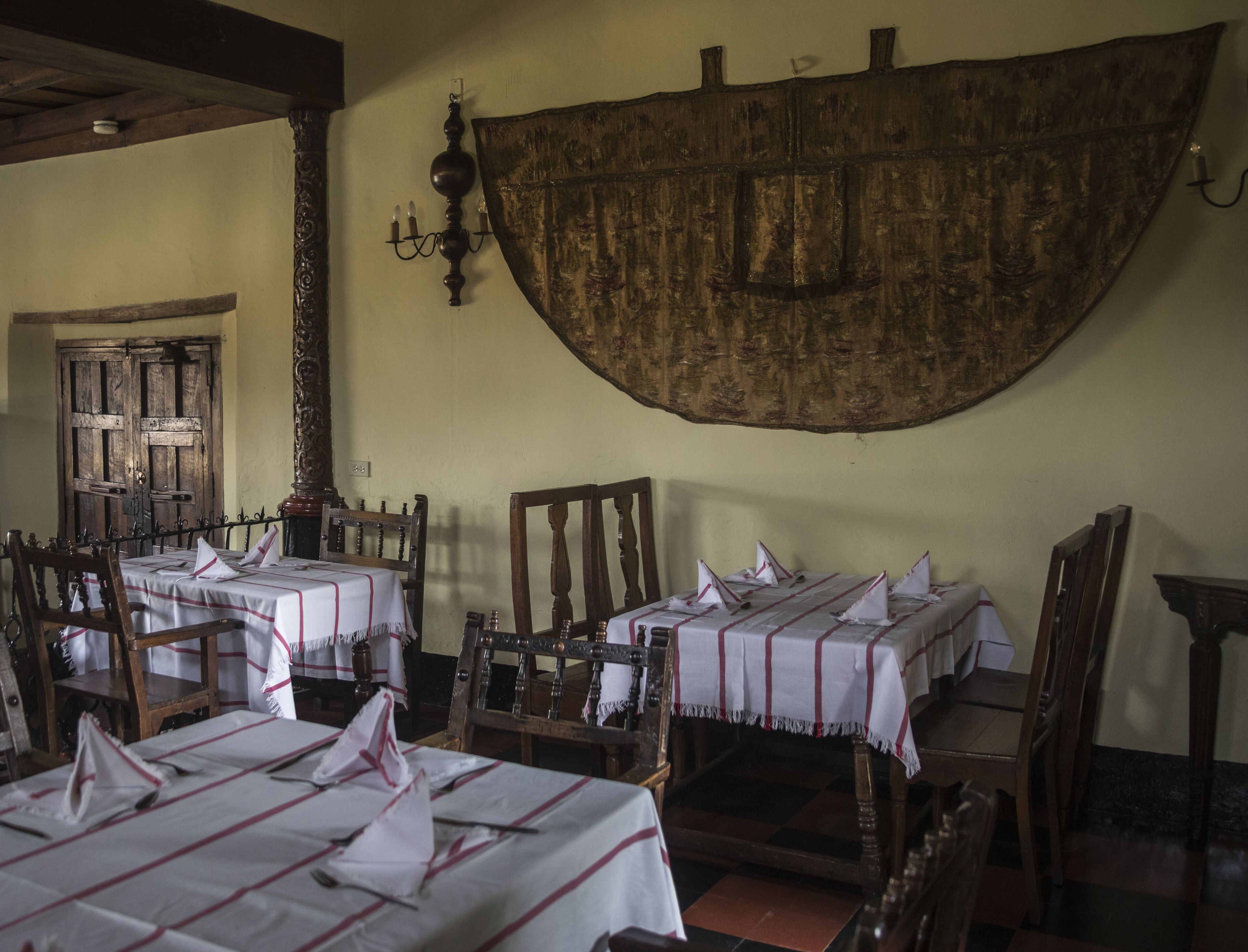 A dining area with tables set for service, wooden chairs, and a large decorative wall piece.