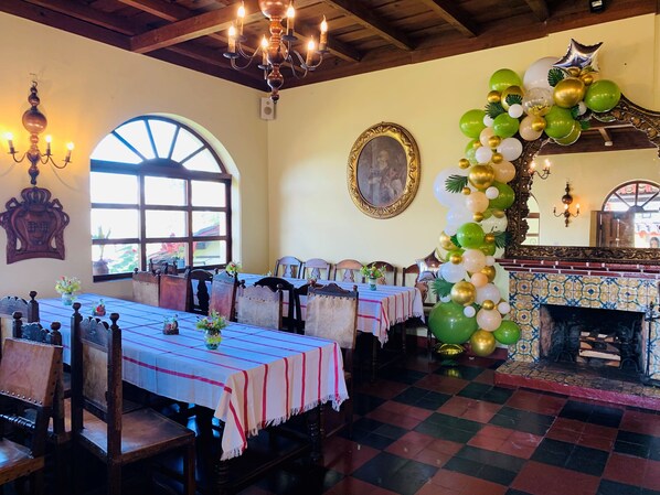 A dining area with a long table, chairs, a chandelier, a fireplace, and a large window.
