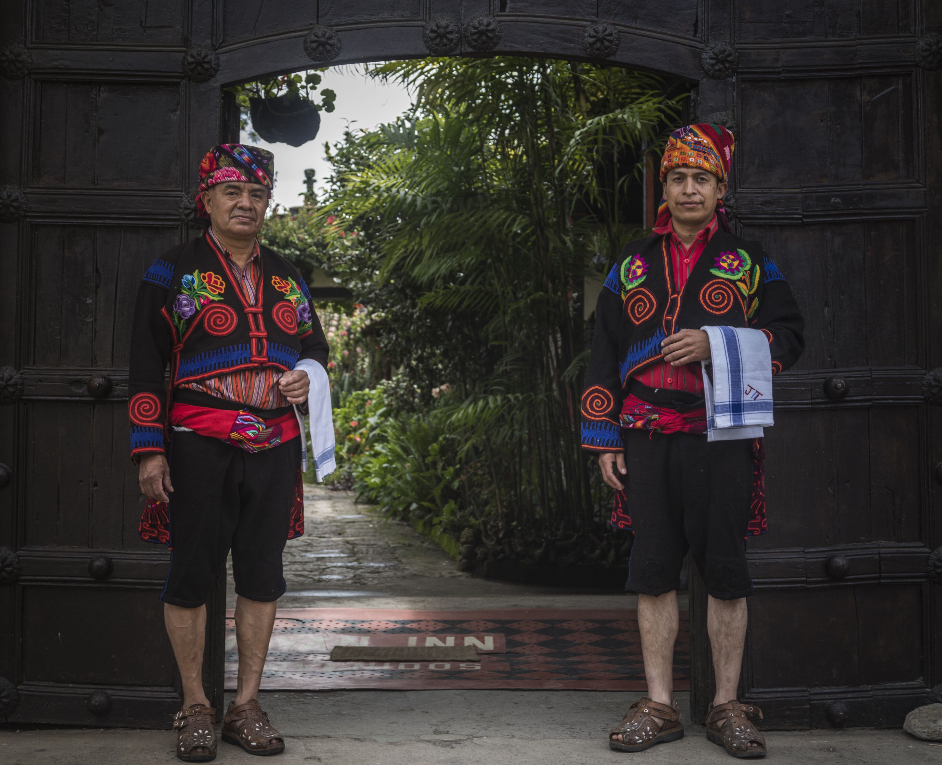 Two individuals in traditional attire standing in front of a wooden archway with a pathway leading through it.