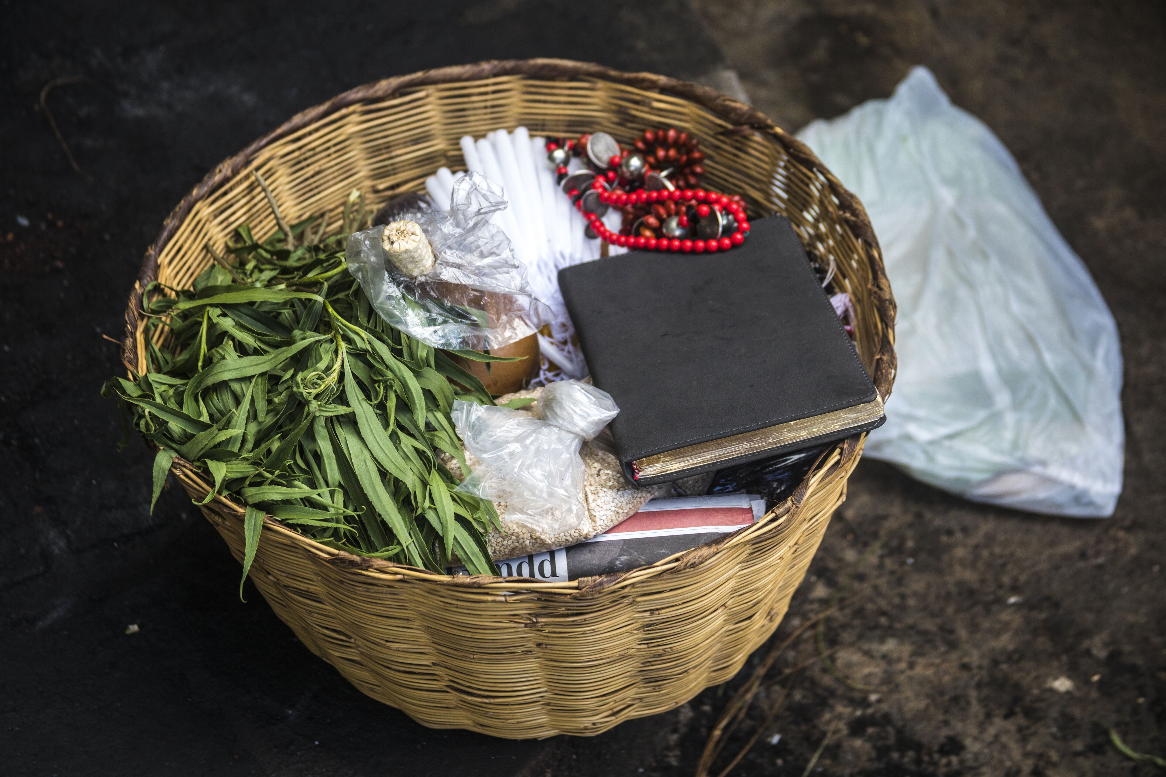 A wicker basket containing green leaves, a plastic bag, a black book, and a red beaded necklace on a dark surface.
